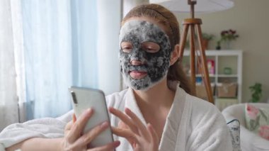 Beyaz bornozlu, derin, saflaştırıcı siyah bir maske takan bir kadın evinde rahat, modern bir iç mekanda akıllı telefonunu kullanıyor..