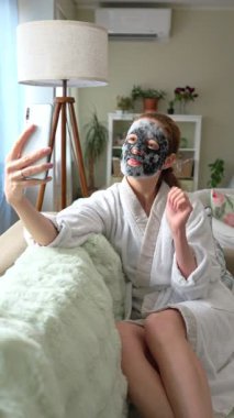 Beyaz bornozlu bir kadın rahat bir koltukta dinleniyor, derin, arındırıcı siyah bir maske takıyor ve akıllı telefonuyla selfie çekiyor..