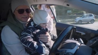 Bir baba ve küçük çocuğu bir arabanın ön koltuğunda oturur ve çocuk heyecanla direksiyona tutunur. Baba onları yakından izliyor, güneş ışığı ön camdan içeri akıyor.