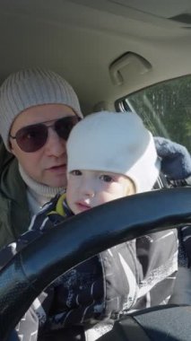 Bir baba ve küçük çocuğu bir arabanın ön koltuğunda oturur ve çocuk heyecanla direksiyona tutunur. Baba onları yakından izliyor, güneş ışığı ön camdan içeri akıyor.