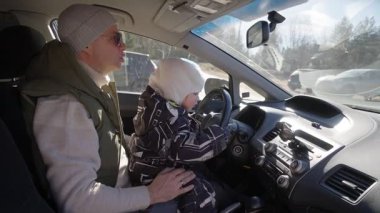 Bir baba ve küçük çocuğu bir arabanın ön koltuğunda oturur ve çocuk heyecanla direksiyona tutunur. Baba onları yakından izliyor, güneş ışığı ön camdan içeri akıyor.