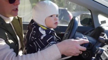 Bir baba ve küçük çocuğu bir arabanın ön koltuğunda oturur ve çocuk heyecanla direksiyona tutunur. Baba onları yakından izliyor, güneş ışığı ön camdan içeri akıyor.