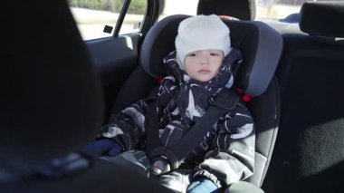 Küçük bir çocuk, sıcak bir kışlık ceket ve beyaz bir şapkayla güvenli bir şekilde bir araba koltuğunda oturuyor. Rahat emniyet kemeri ve rahat kıyafetler güvenliği ve rahatlığı vurguluyor..