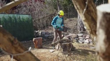 Güçlü, kendine güvenen bir kadın elektrikli testere kullanıyor. Bahar temizliği ve bahçe yenilenmesinin bir parçası olarak eski bir ağacın gövdesini kesiyor. Koruyucu kıyafetler içinde, cinsiyet normlarına meydan okuyor.