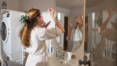 Beyaz bornozlu bir kadın aynaya bakarken anti-reşit bir yüz serumu kullanıyor. Yumuşak doğal ışık, cilt bakımı, güzellik ve kendine özen..