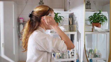 Beyaz bornozlu bir kadın reşit olmayan bir yüz serumu uyguluyor ve aynaya bakarken mikronodik dermaroller kullanıyor. Yumuşak doğal ışık, cilt bakımı, güzellik ve kendine özen..