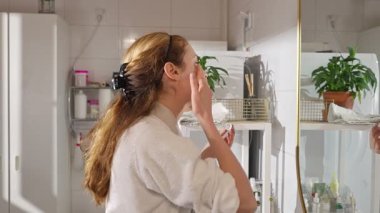 Beyaz bornozlu bir kadın reşit olmayan bir yüz serumu uyguluyor ve aynaya bakarken mikronodik dermaroller kullanıyor. Yumuşak doğal ışık, cilt bakımı, güzellik ve kendine özen..