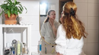 Beyaz bornozlu bir kadın reşit olmayan bir yüz serumu uyguluyor ve aynaya bakarken mikronodik dermaroller kullanıyor. Yumuşak doğal ışık, cilt bakımı, güzellik ve kendine özen..