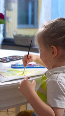 Neşeli genç bir çocuk evde parlak suluboya boyalarla resim yapmaktan zevk alır. Odaklanmış ve mutlu bir şekilde renkli bir sanat eseri yaratıyor. Samimi iç mekan, yumuşak ışıklandırma ve yaratıcı atmosfer bir anı ön plana çıkarıyor.