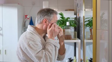 Bornoz giyen olgun bir adam banyo aynasının önünde göz altı bantları takar. Rahat bir ev ortamında sabah rutini, sağIık, kendine dikkat, sağlıklı yaşam tarzı ve erkekler için yaşlanma karşıtı bakım..