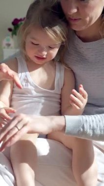 Anne, çocuğun ellerine ve ayaklarına doğal ışık altında nemlendirici krem sürüyor. Nazik cilt bakımı, ebeveyn-çocuk bağı ve sağlıklı cilt koruması. Yakın çekim, sıcak atmosfer, kişisel bakım.