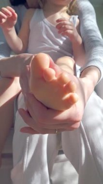 Anne, çocuğun ellerine ve ayaklarına doğal ışık altında nemlendirici krem sürüyor. Nazik cilt bakımı, ebeveyn-çocuk bağı ve sağlıklı cilt koruması. Yakın çekim, sıcak atmosfer, kişisel bakım.