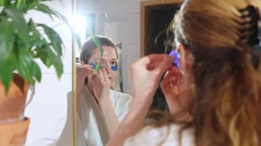 Banyodaki aynadaki yansımasına bakarken göz altı bantları takan bornoz giyen genç bir kadın. Sabah güzellik rutini cilt bakımı ve kişisel bakım için.