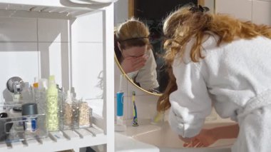 Yüzünü banyo lavabosunda yıkayan, bornoz giyen, aynaya yansıyan, ev bitkileri, havlular ve konforlu ev ortamında çamaşır makinesi olan bir kadın..