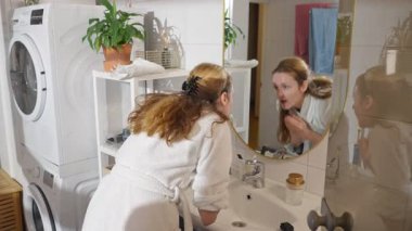 Yüzünü banyo lavabosunda yıkayan, bornoz giyen, aynaya yansıyan, ev bitkileri, havlular ve konforlu ev ortamında çamaşır makinesi olan bir kadın..