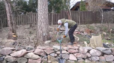 Adam arka bahçedeki çalıları ve küçük ağaçları manuel bahçe makası ile kesiyor. Bahar temizliği ve bahçe işleri, eldivenler ve açık hava kıyafetleri..