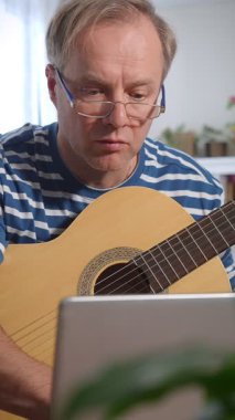 Orta yaşlı bir adam rahat bir odada oturuyor ve akustik gitar çalmayı öğrenmeye odaklanıyor. Önündeki küçük bir masaya yerleştirilmiş dizüstü bilgisayarında bir video dersi izliyor. Kendi kendini öğrenme ve kişisel gelişim anı.