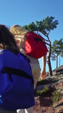 Yaz doğasında yürüyüş yapan iki kadın, biri diğerine kayalık yamaçta yardım ediyor. Açık mavi gökyüzü altında takım çalışması, arkadaşlık ve açık hava macerası.