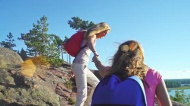 Yaz doğasında yürüyüş yapan iki kadın, biri diğerine kayalık yamaçta yardım ediyor. Açık mavi gökyüzü altında takım çalışması, arkadaşlık ve açık hava macerası.