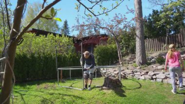 Güneşli bir bahar gününde iki çocuklu bir ailenin arka bahçede trambolin montajı. Açık hava aktivitesi, takım çalışması, ev bahçesi sahnesi..