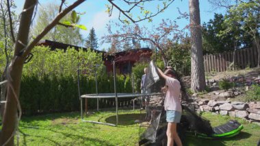 Güneşli bahar gününde aile trambolin topluyor. İki çocuklu baba çerçeveyi hazırlıyor. Açık hava aktivitesi, takım çalışması, ev bahçesi sahnesi..