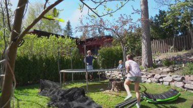 Güneşli bahar gününde aile trambolin topluyor. İki çocuklu baba çerçeveyi hazırlıyor. Açık hava aktivitesi, takım çalışması, ev bahçesi sahnesi..