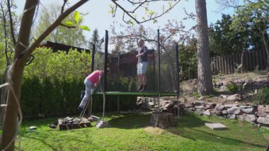 Yetişkin bir adam ve kadın güneşli bir günde arka bahçelerinde trambolinde neşeyle zıplıyorlar. Aktif boş zaman, açık hava eğlencesi, spor ve bahçede kaygısız anlar..