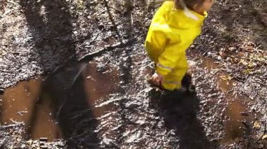 Güneşli bir günde parktaki su birikintisine atlayan sarı yağmurluk giymiş mutlu çocuk. İskandinav ormanlık alanında aktif oyun, eğlence ve doğa keşfi.