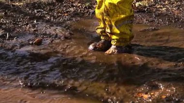 Güneşli bir günde parktaki su birikintisine atlayan sarı yağmurluk giymiş mutlu çocuk. İskandinav ormanlık alanında aktif oyun, eğlence ve doğa keşfi.