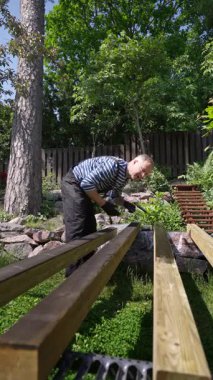 Güneşli bir günde ahşap kirişleri paten fırçasıyla dışarıda boyayan bir adam. Orta yaşlı erkek, çizgili gömlek giyiyor, iş tulumu ve eldiven giyiyor, yeşil bir bahçede çalışıyor. DIY arka bahçe marangozluk projesi