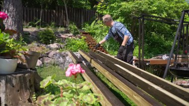 Güneşli bir günde ahşap kirişleri paten fırçasıyla dışarıda boyayan bir adam. Orta yaşlı erkek, çizgili gömlek giyiyor, iş tulumu ve eldiven giyiyor, yeşil bir bahçede çalışıyor. DIY arka bahçe marangozluk projesi