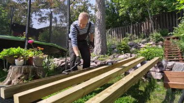 Güneşli bir günde ahşap kirişleri paten fırçasıyla dışarıda boyayan bir adam. Orta yaşlı erkek, çizgili gömlek giyiyor, iş tulumu ve eldiven giyiyor, yeşil bir bahçede çalışıyor. DIY arka bahçe marangozluk projesi