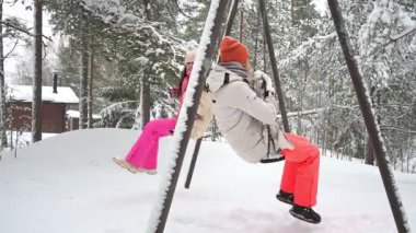 Middle-aged mother and teenage daughter enjoy winter holidays in Finland, swinging together in snowy park. Warm clothes, joyful emotions, family values, seasonal lifestyle and vacation mood.