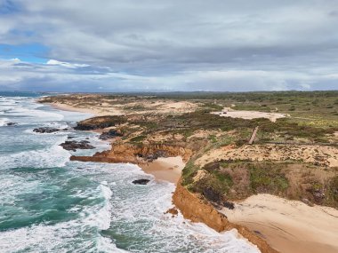 Okyanus kıyısının havadan görünüşü, yüksek uçurumlar, çarpan dalgalar ve baharda balıkçıların patikası boyunca kum tepeleri. Alentejo, Portekiz