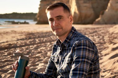 Gün batımında kayalık bir plajda oturan ve elinde termal bardak tutan bir adamın portresi. Bulanık arka plandaki okyanus, kum ve kayalıklar rahat ve sakin bir atmosfer yaratır.