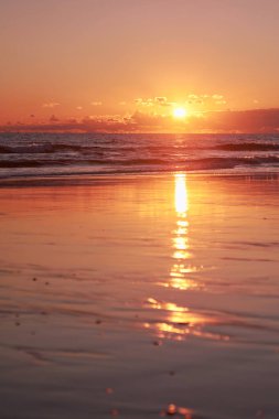 Güneş ufukta görünürken ve gökyüzünde ve suda altın bir parıltıyla okyanusun üzerinde huzurlu bir gün batımı. Islak kum, günbatımının canlı turuncu rengini yansıtır ve aynaya benzer bir etki yaratır.