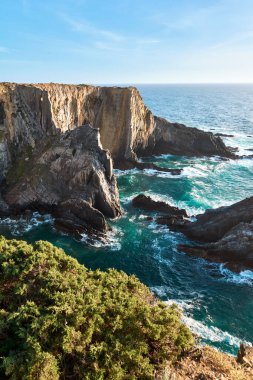 Dik kayalıklarıyla baş döndürücü kıyı manzarası, aşağı düşen turkuaz dalgalar, ve ön planda verimli yeşillik Portekiz Balıkçı Patikası boyunca doğal güzellik ve sükunet yayıyor.