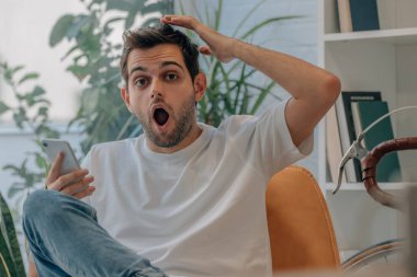 Genç adam akıllı telefonu ve şaşkınlık ifadesiyle evde.