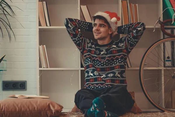 クリスマスのセーターとサンタ クラスの帽子でリラックスした家の若い男 — ストック写真