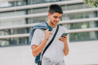 Genç adam sokakta yürüyor ve cep telefonuna bakıyor.