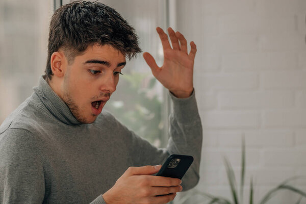 молодой человек дома смотрит на мобильный телефон с удивлением