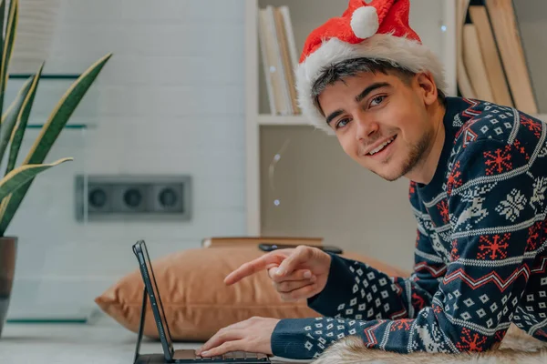 サンタクラスの帽子でクリスマスに家で笑顔ノートパソコンの画面を指している若い男 — ストック写真