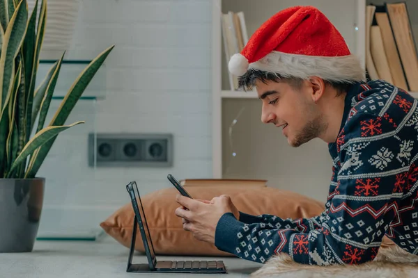 サンタ クラスの帽子をかぶった若い男がクリスマスに自宅でコンピュータと携帯電話を持っています — ストック写真