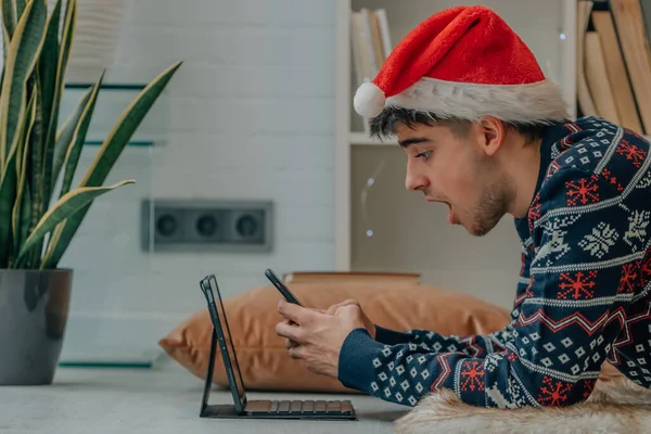 サンタ クラスの帽子と驚きの表情でクリスマスに自宅でコンピュータと携帯電話を持つ若い男 — ストック写真