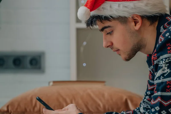 サンタ クラスの帽子を被った若い男が携帯電話を持って家にいて — ストック写真