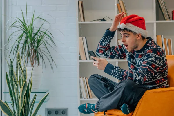 クリスマスに携帯電話を持って家にいる若い男と驚きの表情 — ストック写真