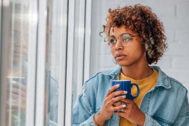 Evdeki düşünceli kadın elinde fincanla pencereden dışarı bakıyor.