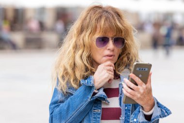 middle-aged woman with mobile phone in the street with a thoughtful or distrustful expression of doubt
