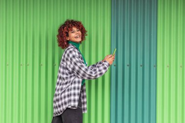 Cep telefonu olan bir kız yeşil duvarla sokakta yürüyor.