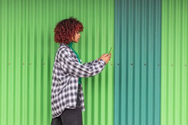 Cep telefonu olan bir kız yeşil duvarla sokakta yürüyor.